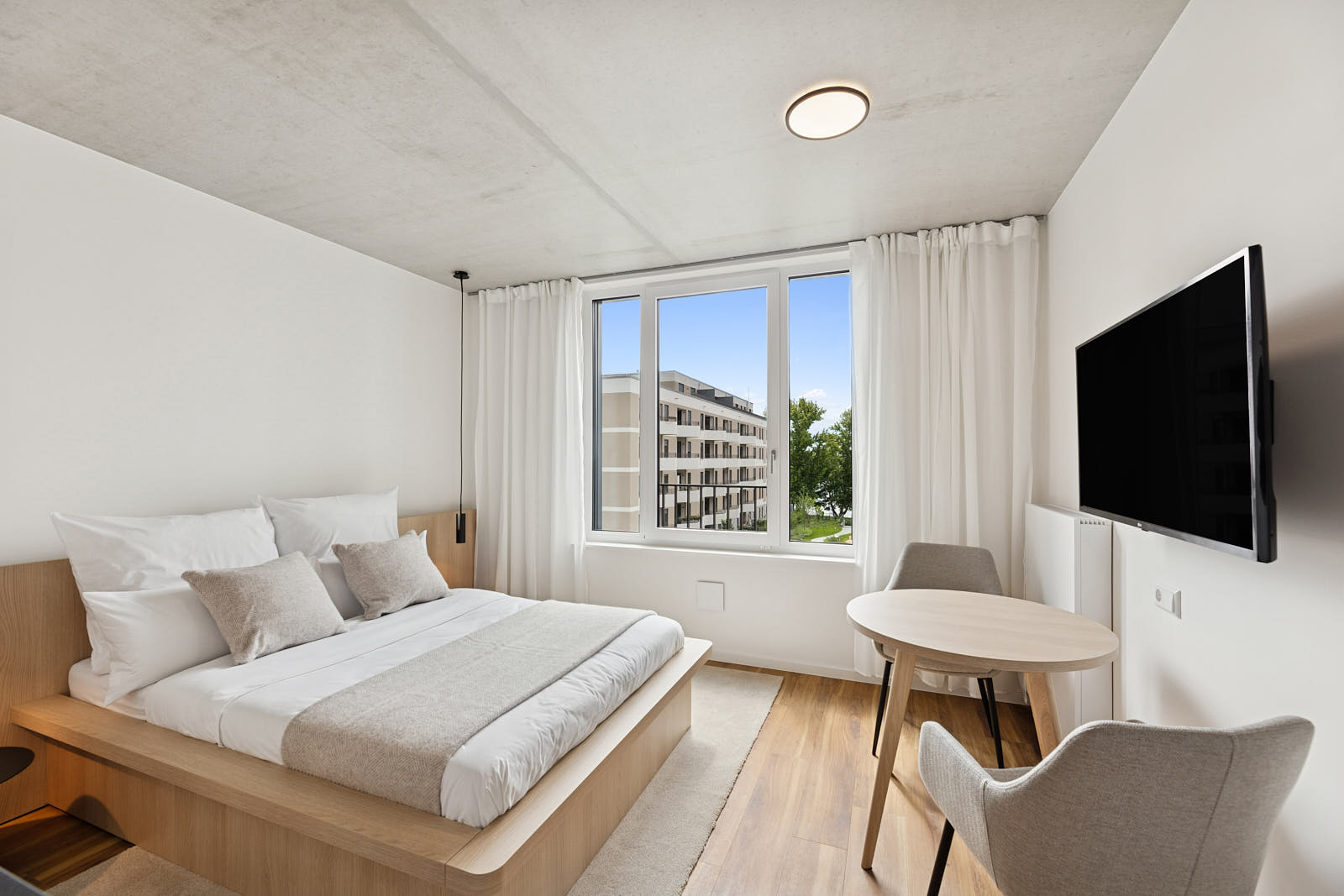 Modern eingerichtetes Schlafzimmer mit großem Fenster, Sitzbereich und Blick auf ein Wohngebäude
