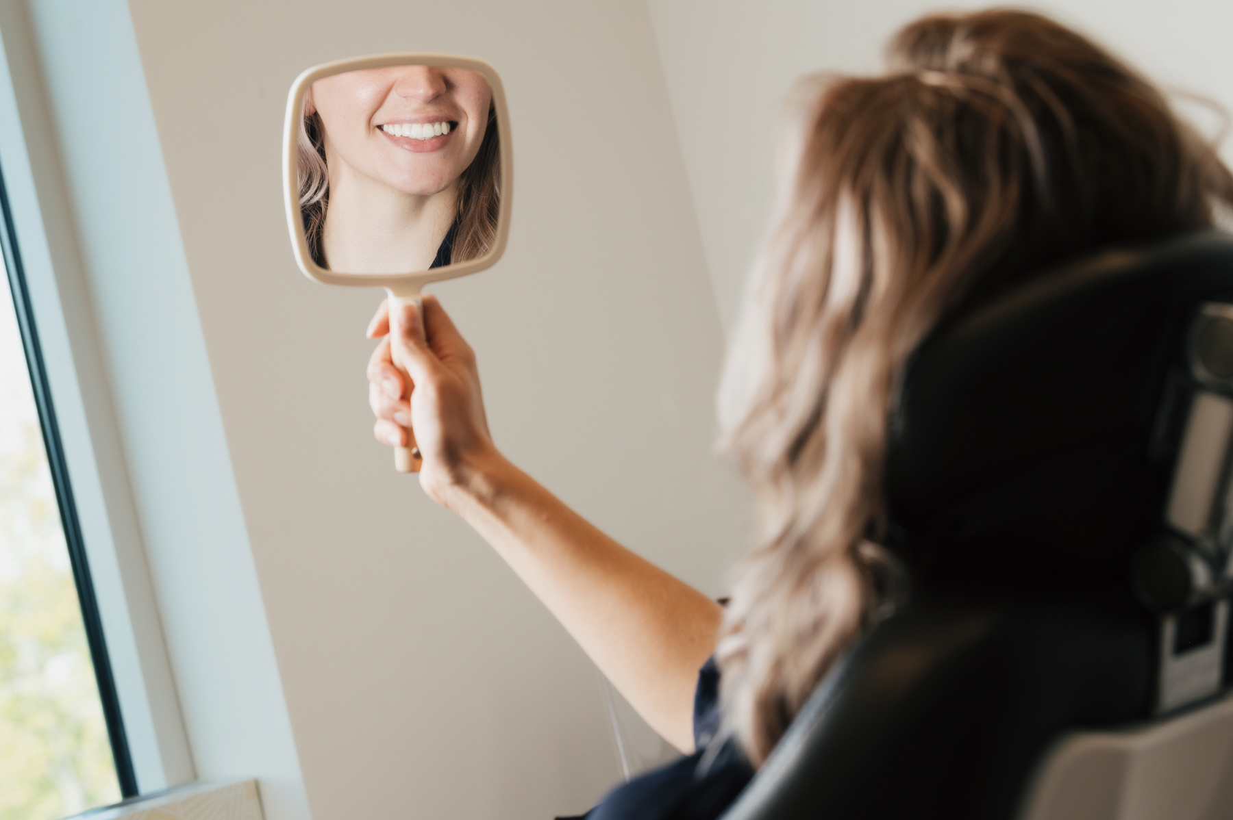 Woman with wavy hair sitting in a chair holding a hand mirror reflecting her smiling mouth and teeth.
