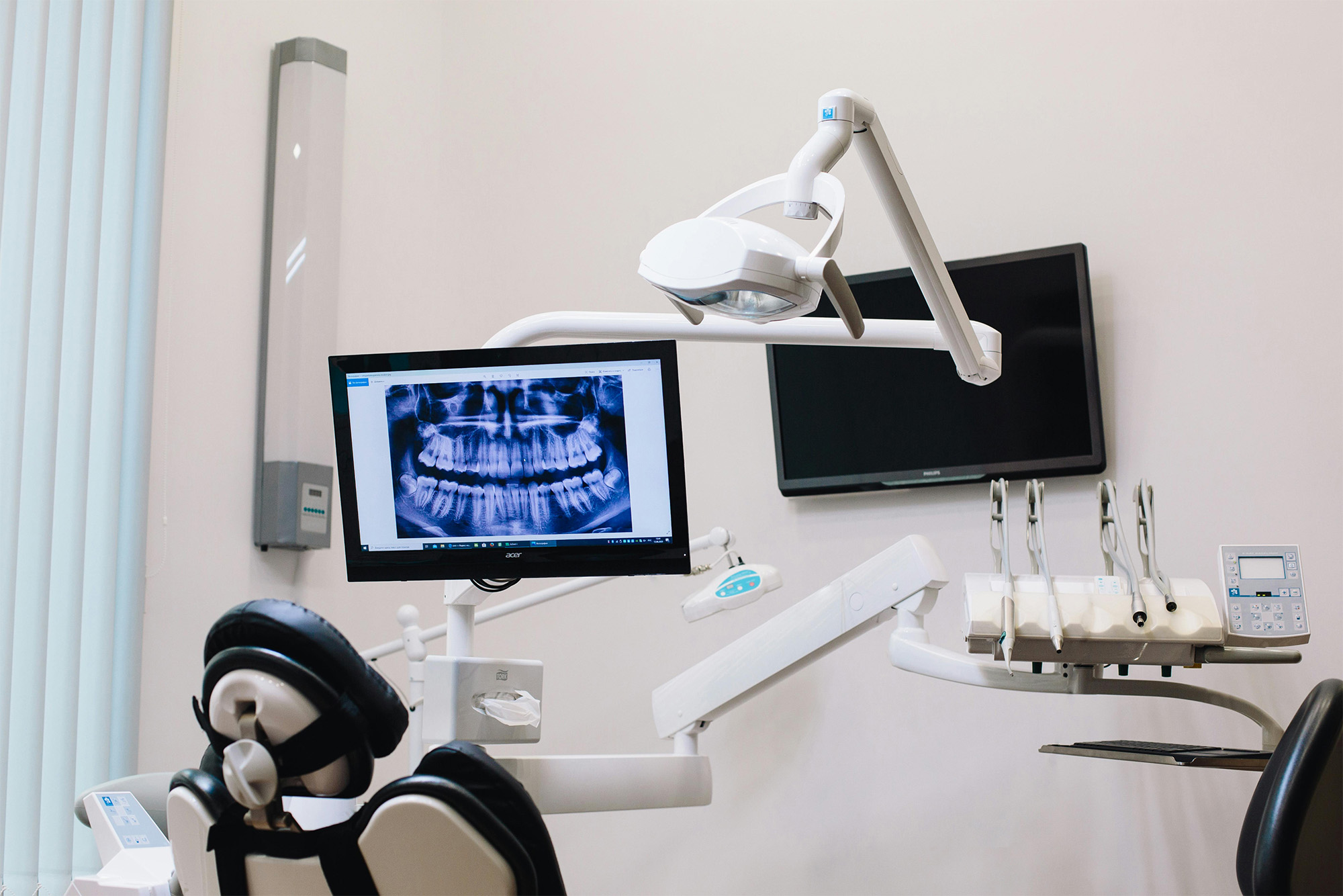 Dental office setup with a patient chair, dental tools, overhead light, and a monitor displaying a dental X-ray image.