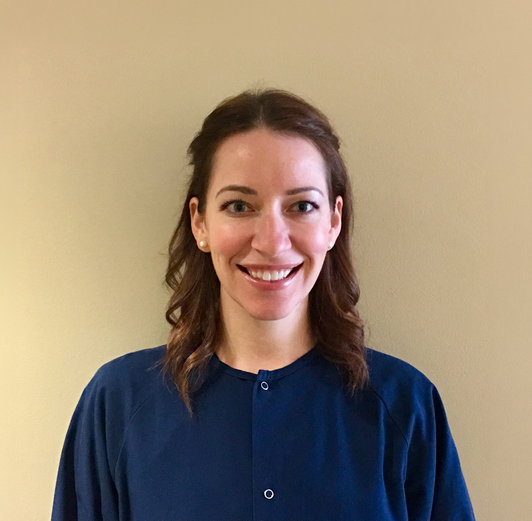 Smiling woman with shoulder-length brown hair wearing a dark blue top against a beige background.