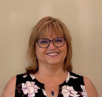 Smiling woman with glasses and light brown hair wearing a black and white floral top against a plain beige background.