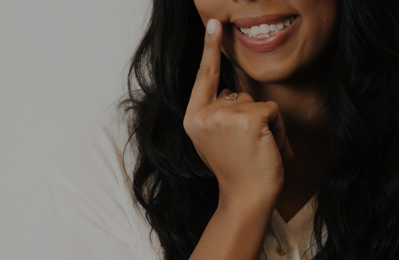 Close-up of a smiling woman with long dark hair pointing to her cheek with her index finger.