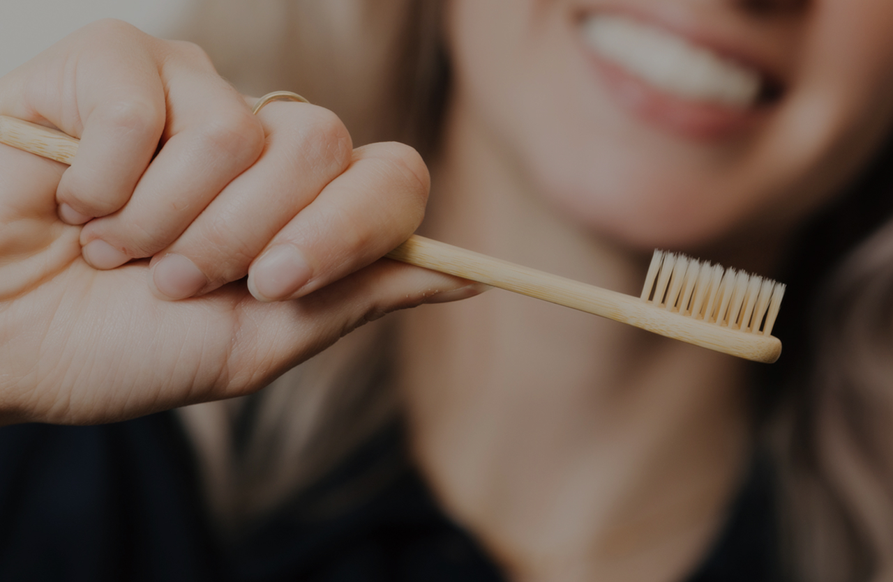 Woman holding a bamboo toothbrush with a blurred smiling face in the background.