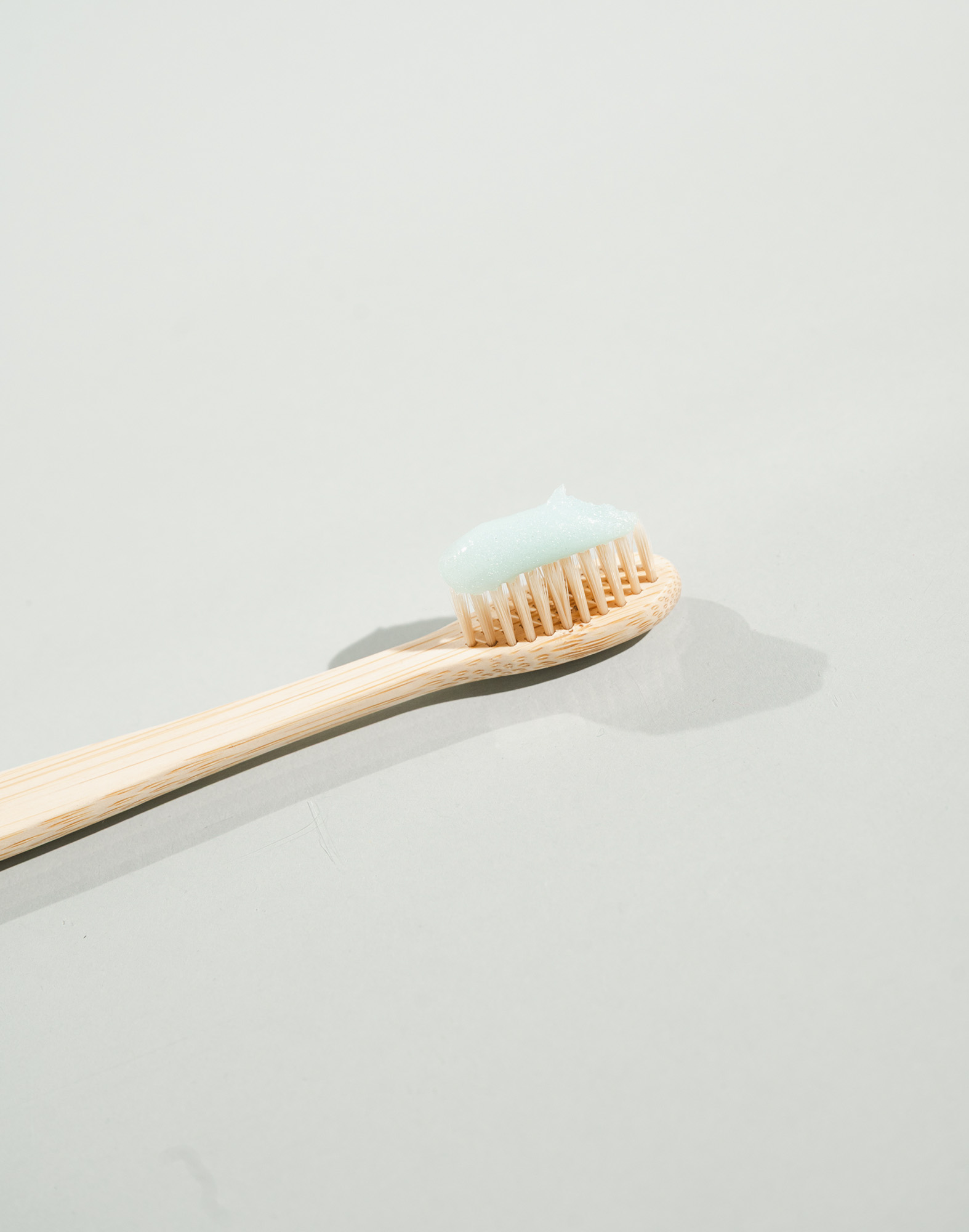 Bamboo toothbrush with light blue toothpaste on bristles against a pale gray background.