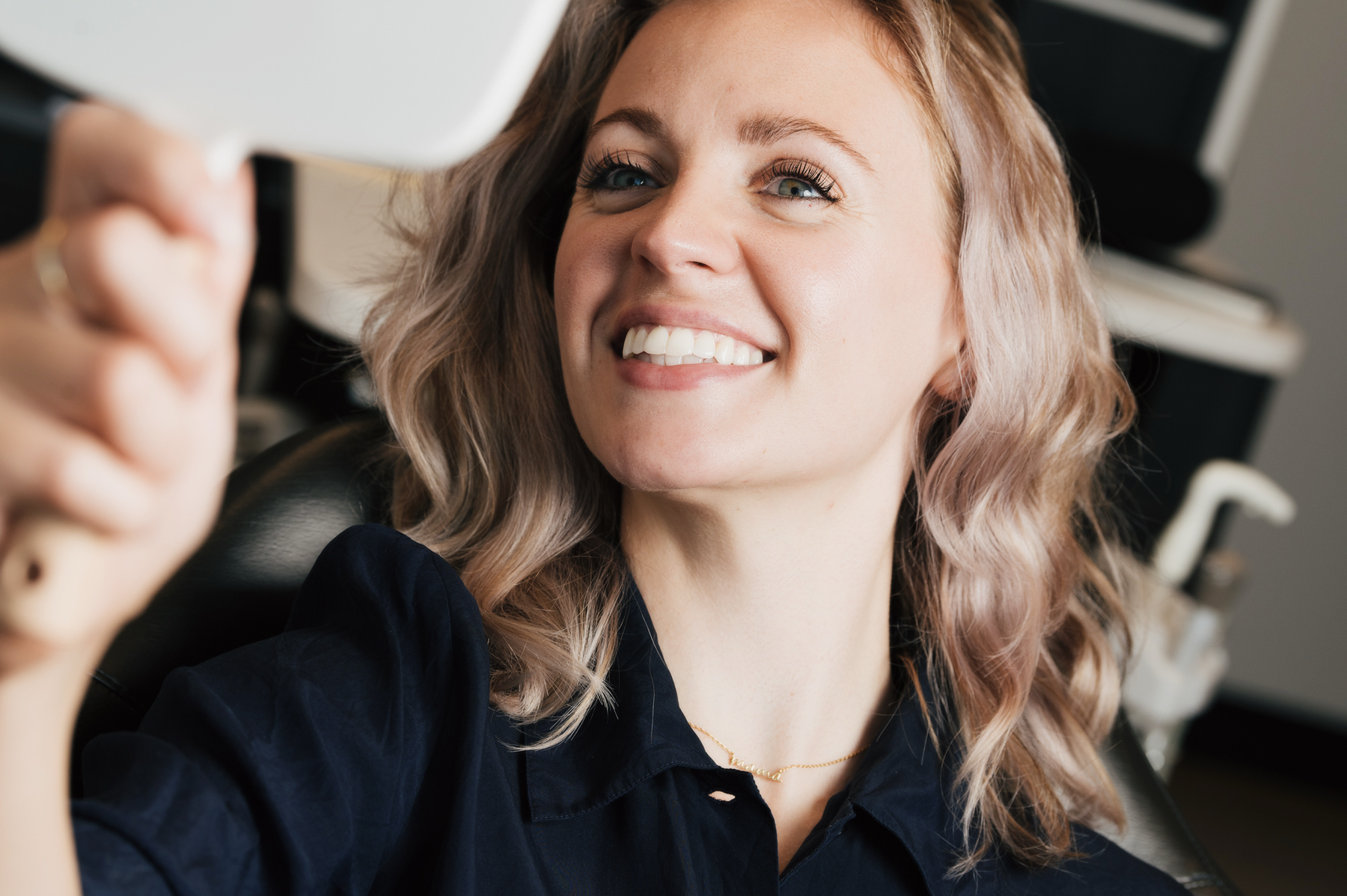Smiling woman with wavy blonde hair looking at her reflection in a handheld mirror.