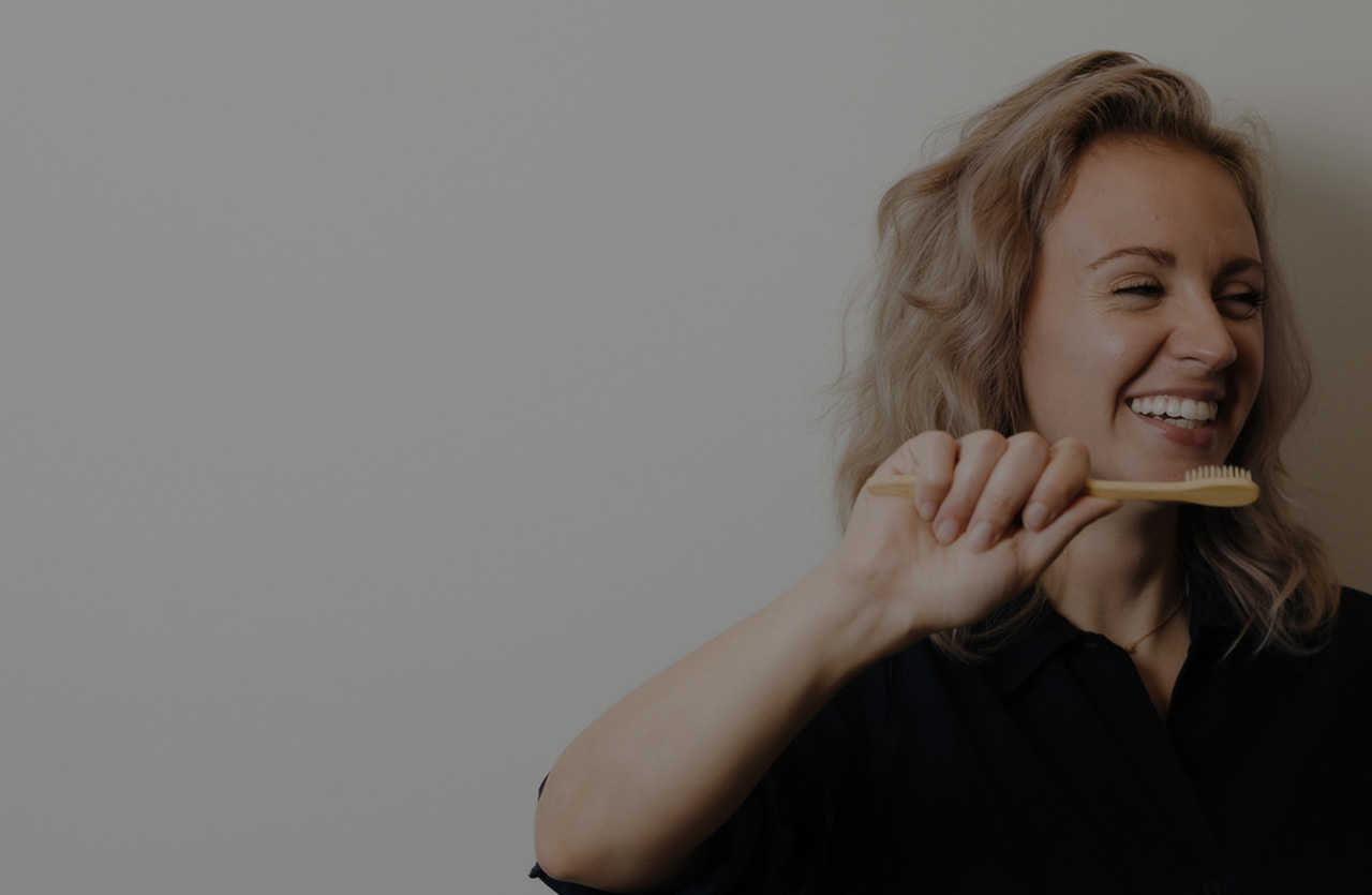 Smiling woman with light curly hair holding a bamboo toothbrush near her mouth.