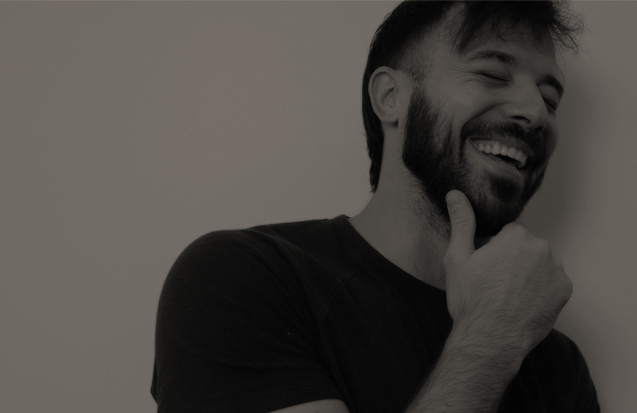 Bearded man smiling with eyes closed and hand touching his chin against a plain background.