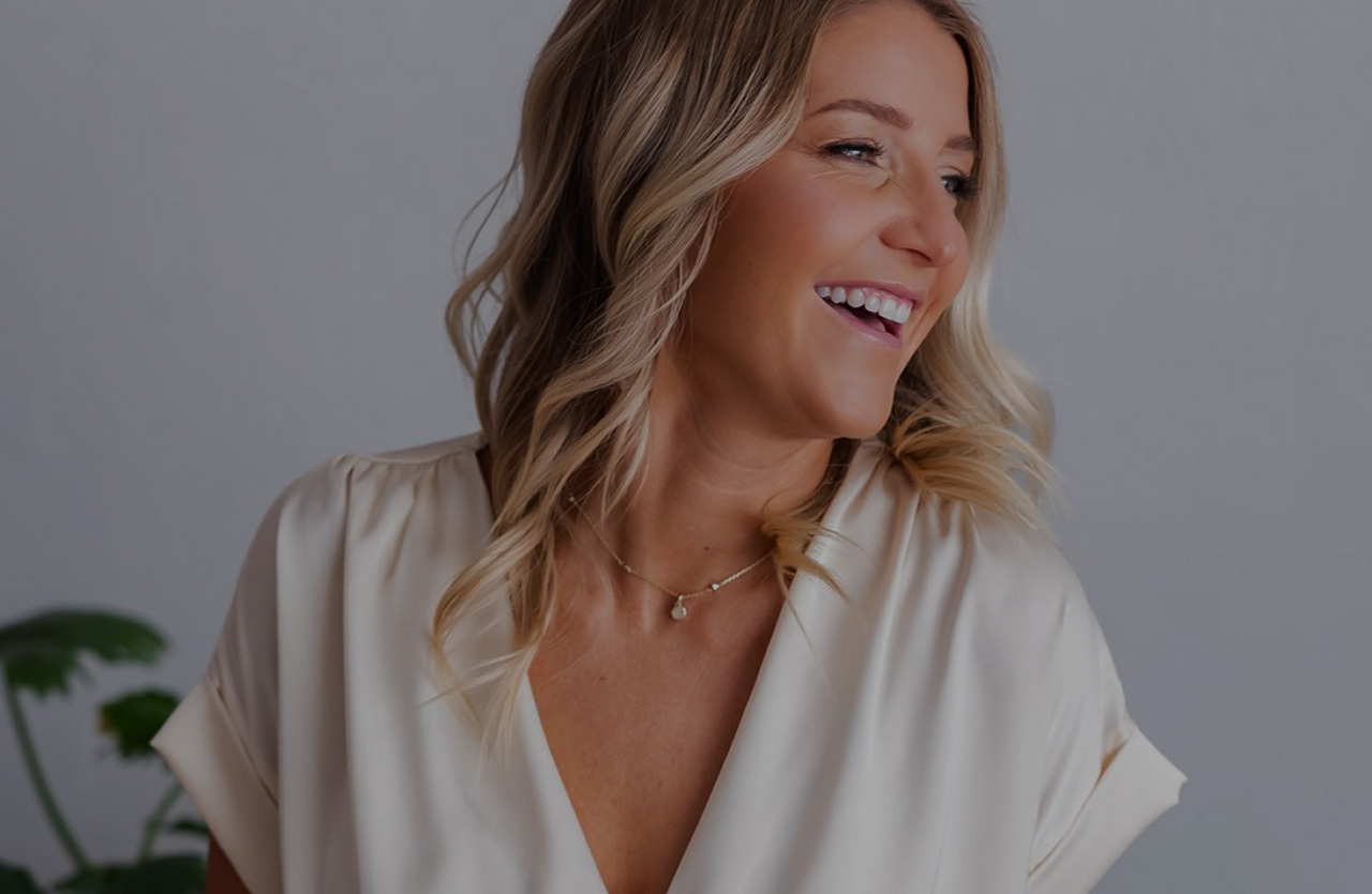 Smiling woman with blonde hair in loose waves wearing a cream blouse and delicate gold necklace against a plain gray background.