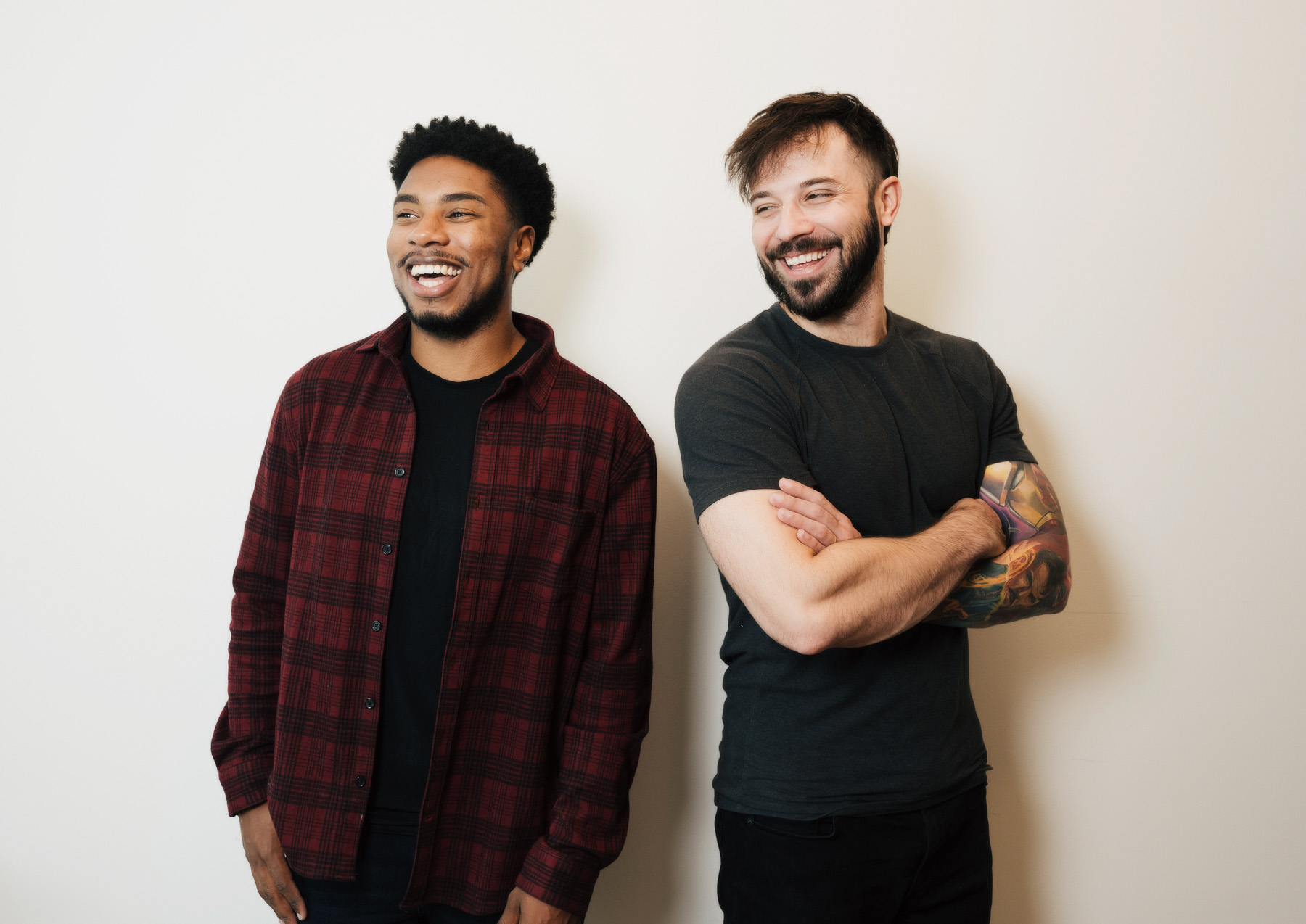 Two men standing against a white wall, smiling and looking to the side; one wears a red plaid shirt and the other has a black t-shirt with a tattooed arm crossed.