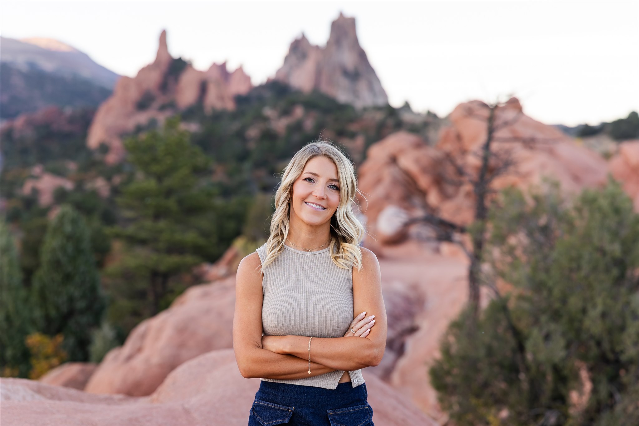 Smiling blonde woman with crossed arms standing outdoors with red rock formations and greenery in the background.