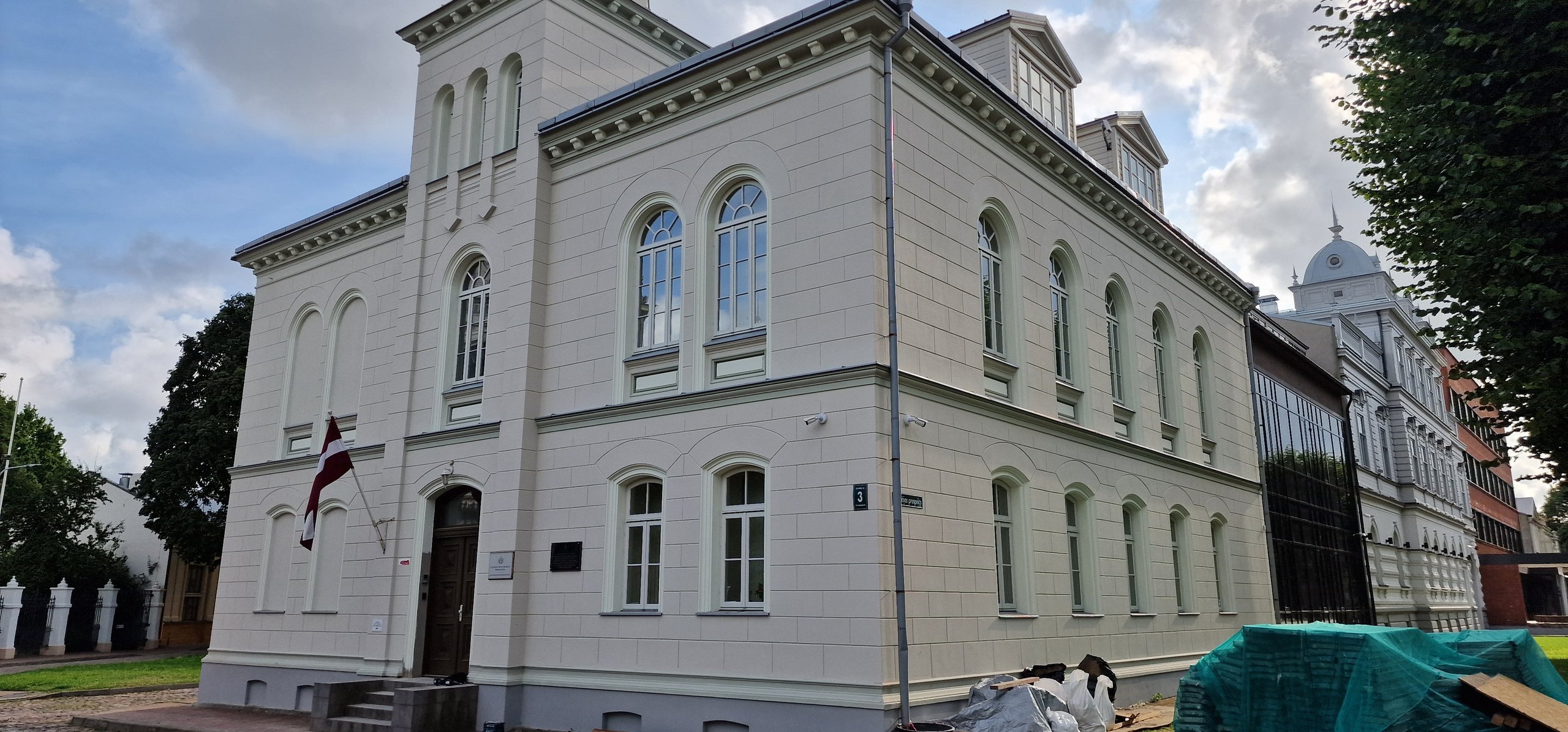 Cream-colored historic building with arched windows and a Latvian flag near the entrance on a partly cloudy day.
