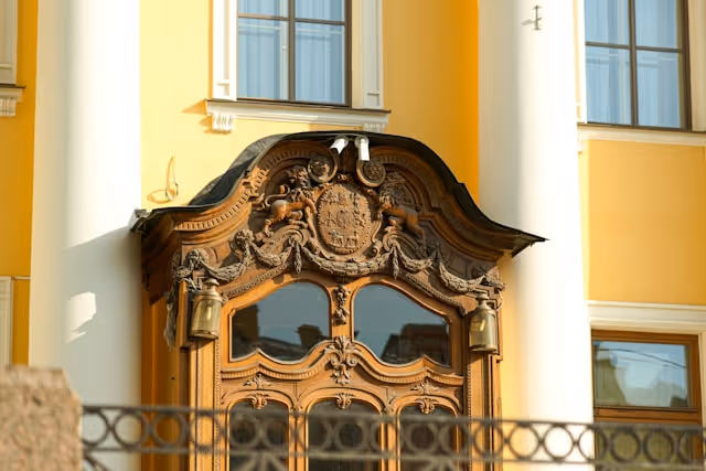 Ornate wooden doorway with intricate carvings and a crest on a yellow building with white columns.