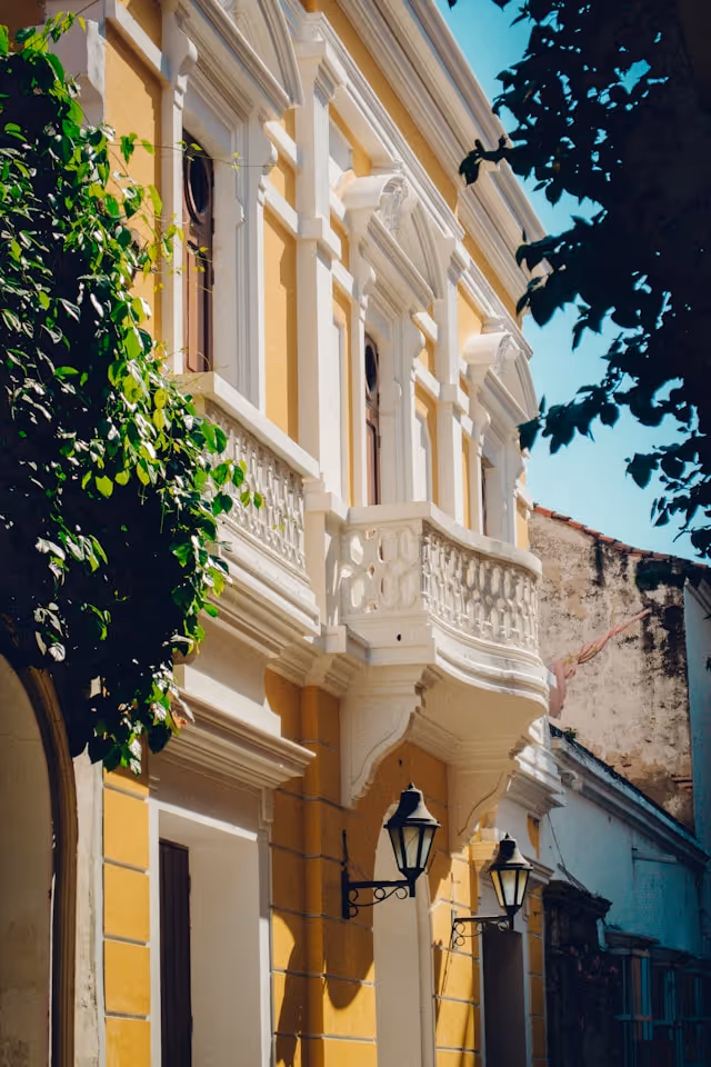 Yellow and white colonial-style building facade with ornate balcony and two black wall-mounted lanterns.