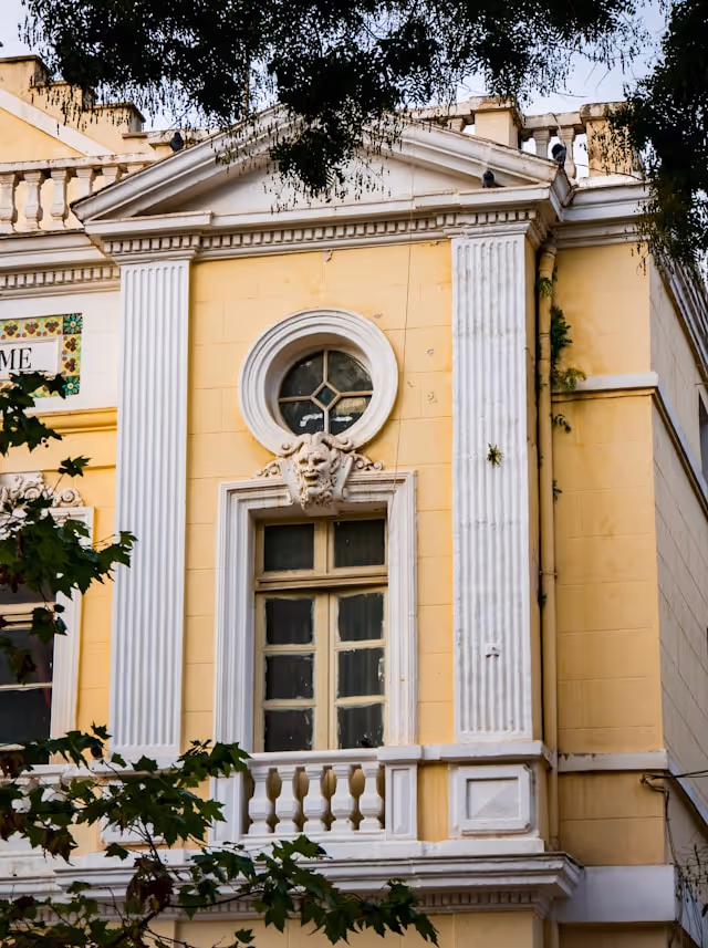 Upper section of a yellow classical building facade with white columns, a circular window, decorative lion head, and leafy tree branches.