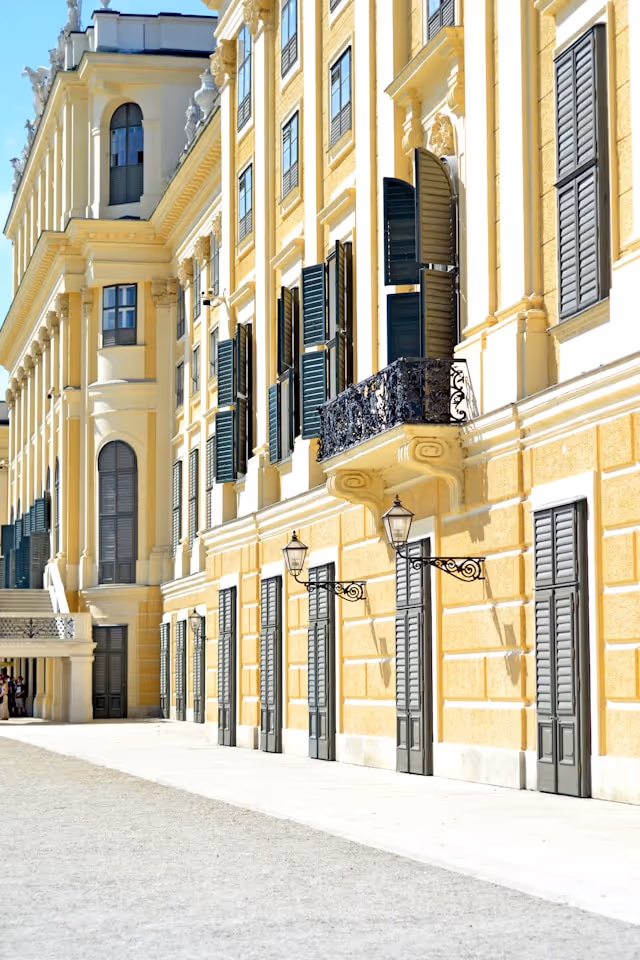 Sunny facade of a classical yellow building with green shutters and ornate black balcony railing.