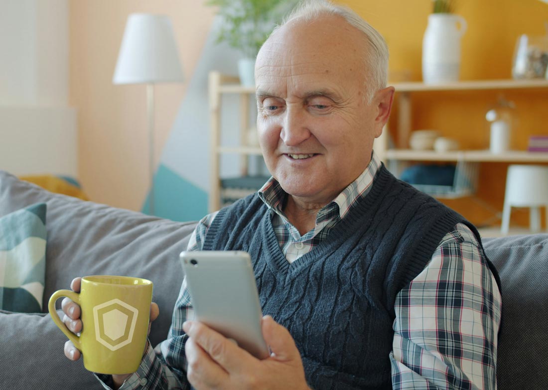 Älterer Mann sitzt mit Kaffeetasse auf Sofa und schaut lächelnd auf sein Smartphone.
