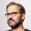 Mature man with glasses and beard looking to the side against a plain background.