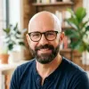 Smiling bald man with glasses and beard wearing a dark blue shirt indoors with plants in background.