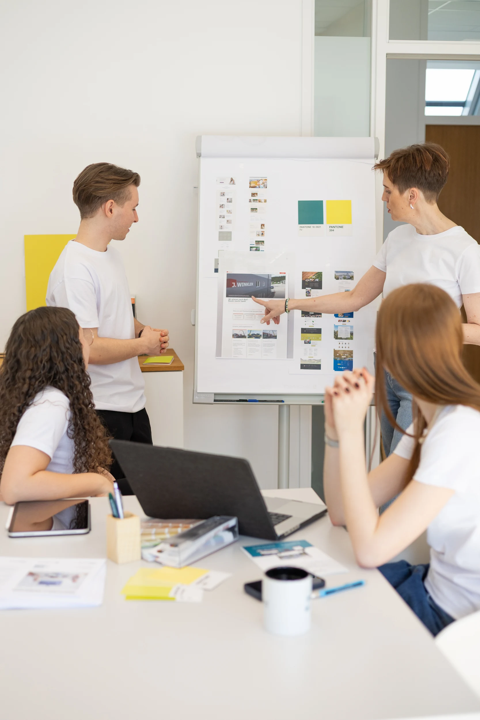 Vier Kollegen in weißen Hemden halten eine Besprechung ab, wobei einer auf ein Flipchart mit Farbmustern und Webdesign-Entwürfen zeigt.