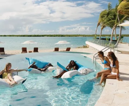 Four women relaxing on inflatable pool floats and poolside by palm trees with ocean in background.
