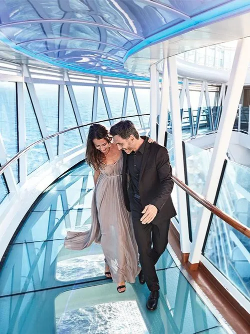 Couple dressed formally walking on a glass-floored corridor on a cruise ship with ocean views.