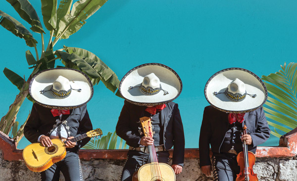 Three mariachi musicians in traditional attire playing guitar, vihuela, and violin outdoors.