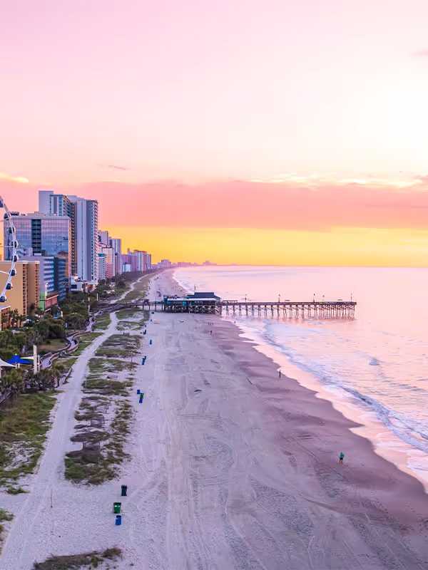 Sunset view of empty beach with pier and city buildings lining the shore under pink and yellow sky.