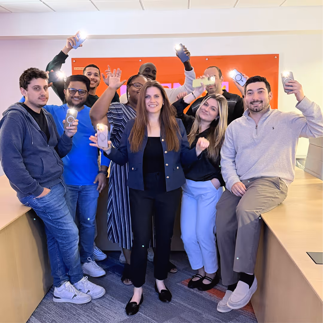 Group of diverse people smiling indoors holding phones with flashlights on, against an orange wall.