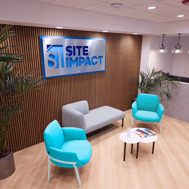 Office lounge with teal chairs, gray sofa, wood panel wall, and Site Impact sign in blue and silver.