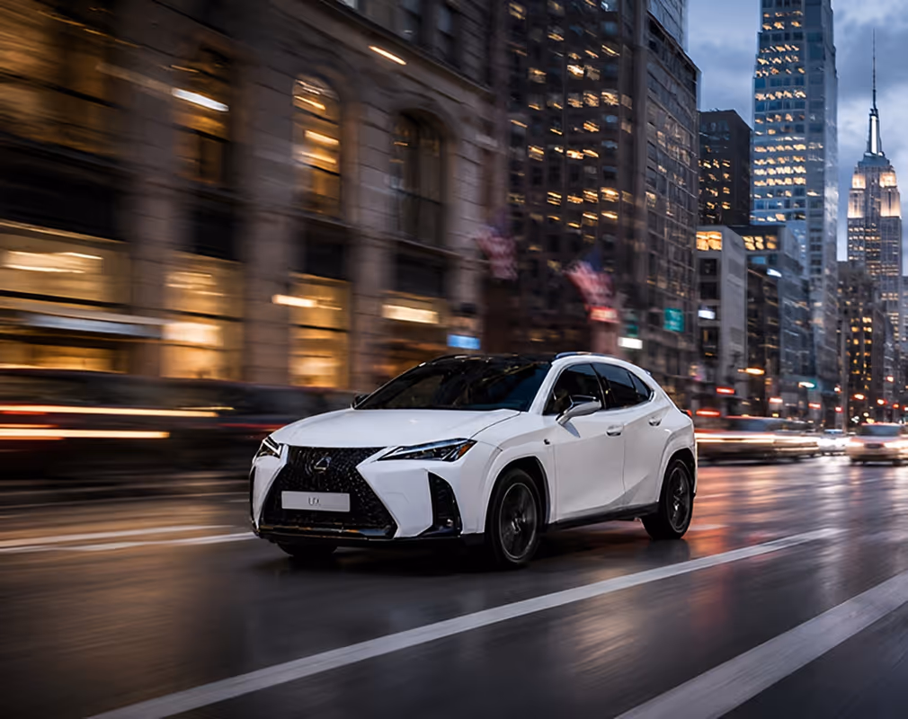White Lexus SUV driving fast on a city street at dusk with blurred lights and buildings in the background.