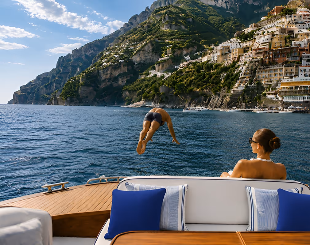 Man diving into sea from a boat with a woman watching and cliffside buildings in the background.
