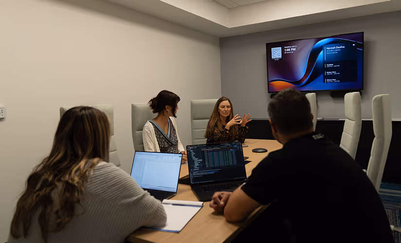 Four people in a meeting room with laptops, discussing while looking at a screen displaying schedule details.