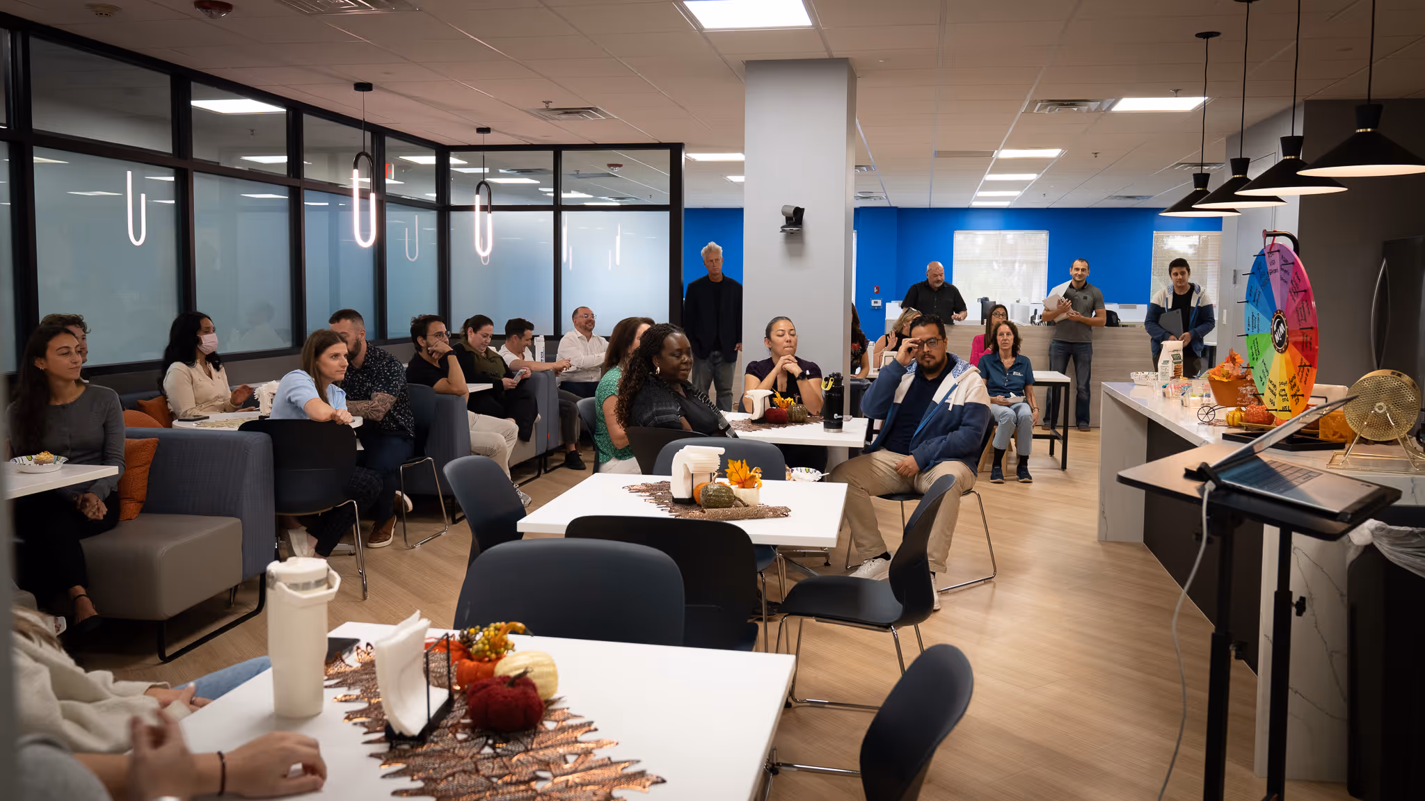 Group of people seated and standing inside a modern office space with tables and a colorful prize wheel.