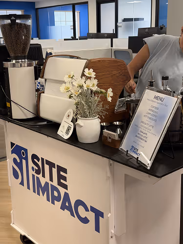Coffee station with espresso machine, menu, flowers, and person preparing a drink in an office setting.