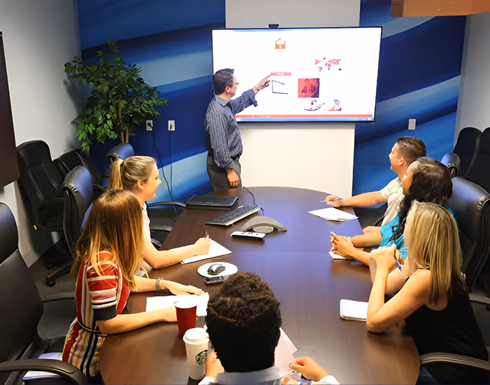 Man presenting data on a screen to six colleagues seated around a conference table in a meeting room.