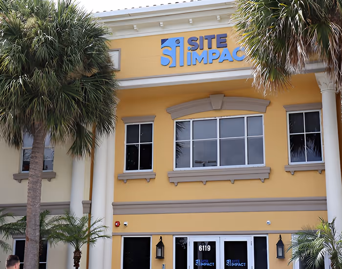 Yellow building with white columns and Site Impact sign above entrance under palm trees.