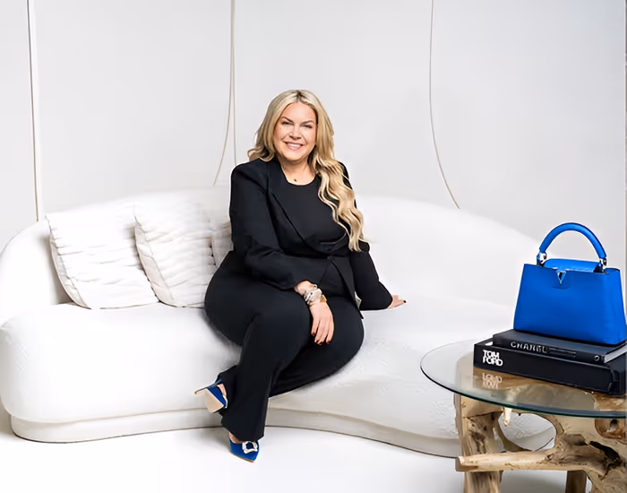 Smiling woman in black suit sitting on white curved sofa with blue handbag on glass table.
