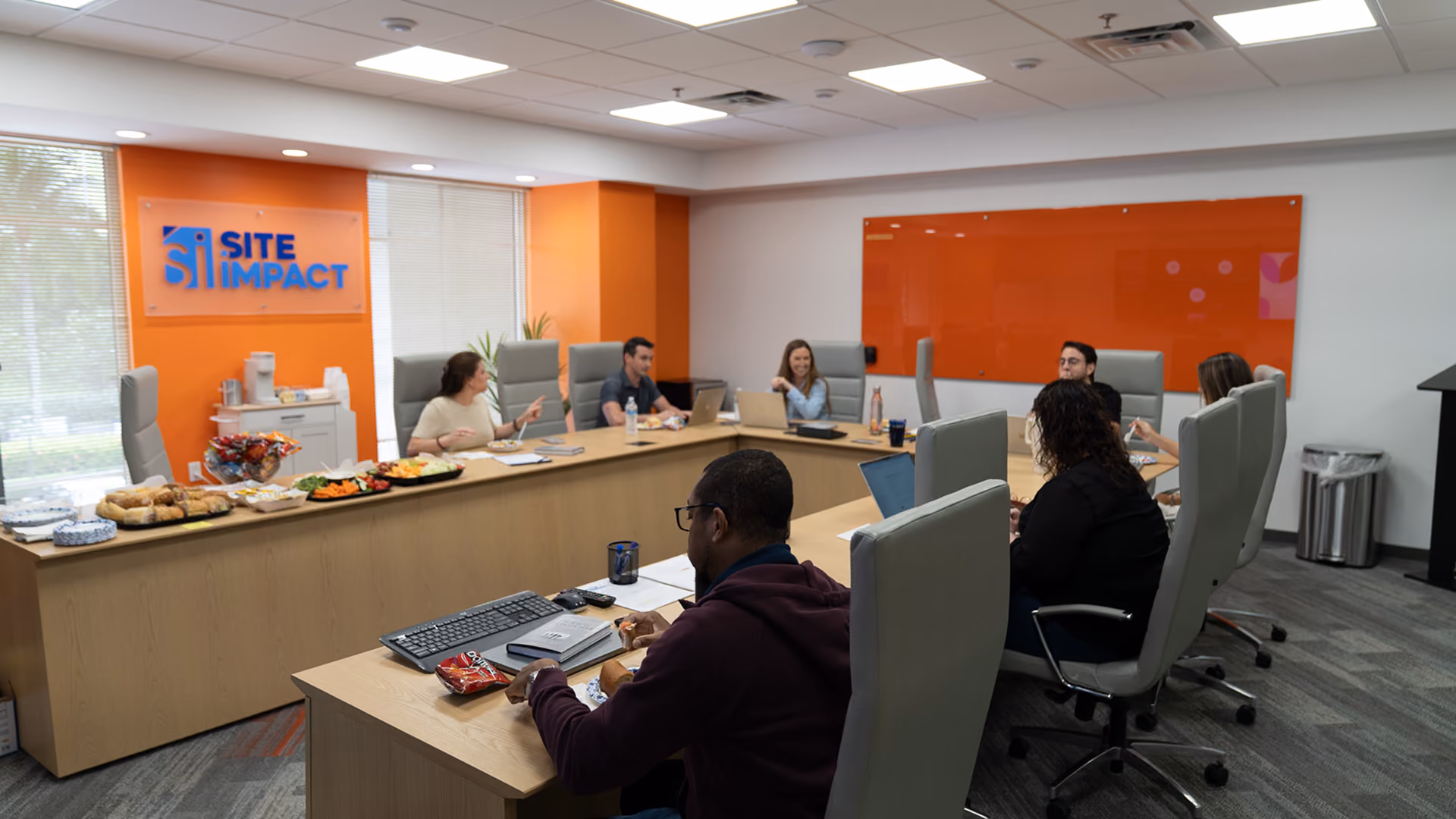 Seven people having a meeting in a conference room with laptops and snacks on the table.