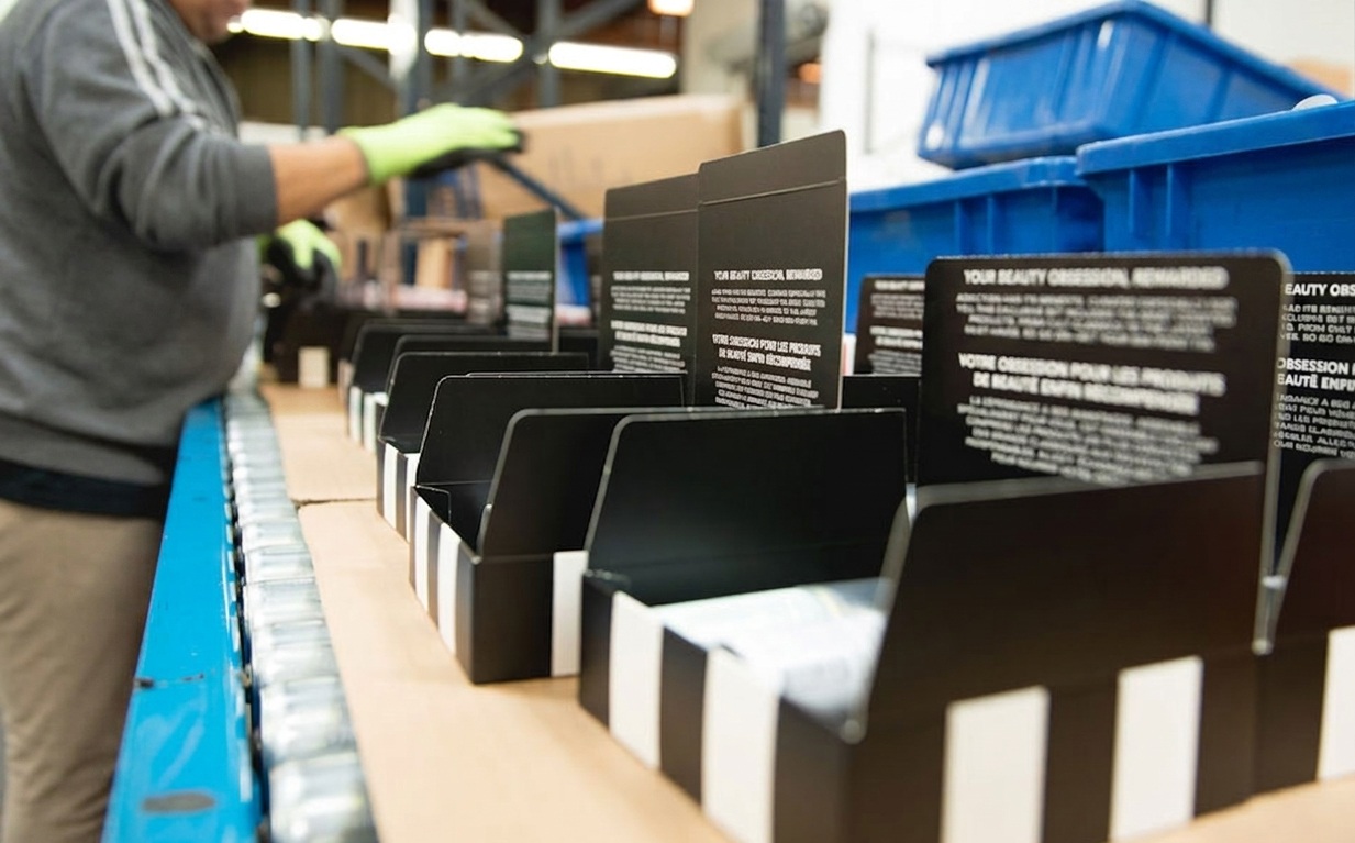 Warehouse assembler building branded beauty subscription kits on a conveyor line, with open black and white striped boxes staged for component insertion at a 3PLCity kitting and assembly station.