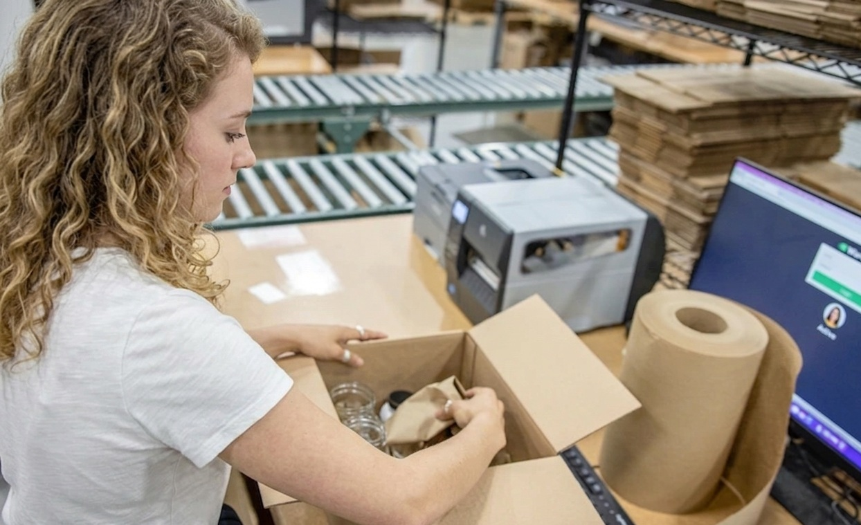 3PLCity packing associate placing supplement and vitamin bottles into a shipping box with kraft paper void fill at a packing station with a label printer, conveyor line, and order screen visible.