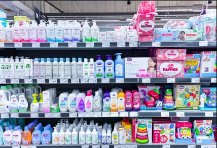 A retail display of baby care essentials organized on shelves. The selection includes several rows of infant shampoos, soaps, and lotions in pump and squeeze bottles, as well as baby wipes, diapers, and a variety of colorful developmental toys like stackin