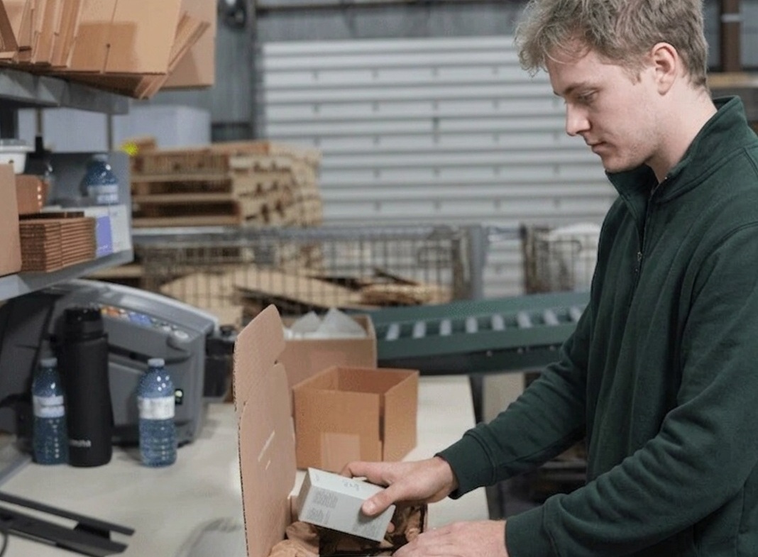 A warehouse employee in a green pullover packing a e-commerce order at a fulfillment station.
