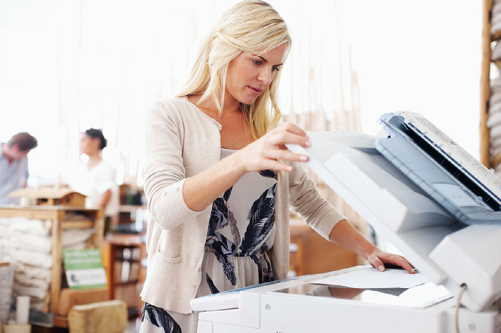 A woman making copies on a multi-function printer. With two people in the back talking about houses