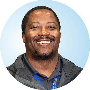 Corey smiling at the camera wearing a dark collared shirt and lanyard against a light blue circular background.