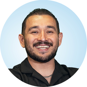 Daniel smiling at the camera with a trimmed beard and black collared shirt against a light blue circular background.
