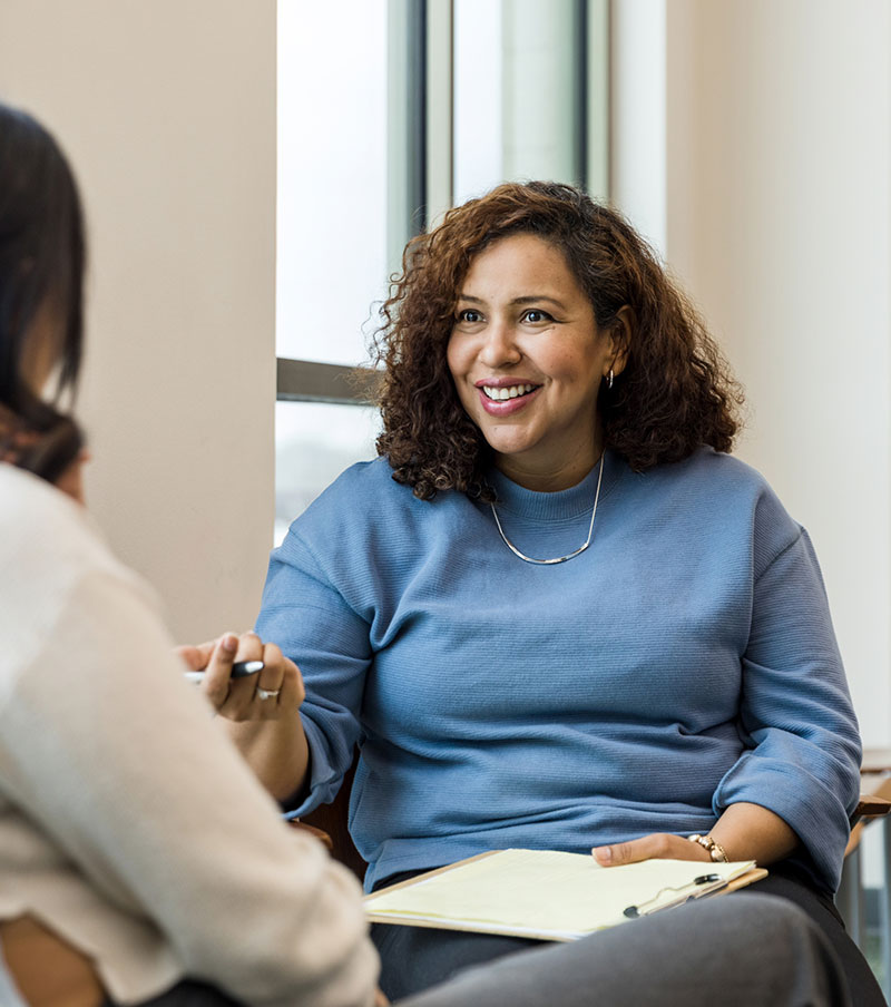 A Tri-City Mental Health employee sits in a counseling setting speaking with a community member while holding a notepad during a supportive conversation.”