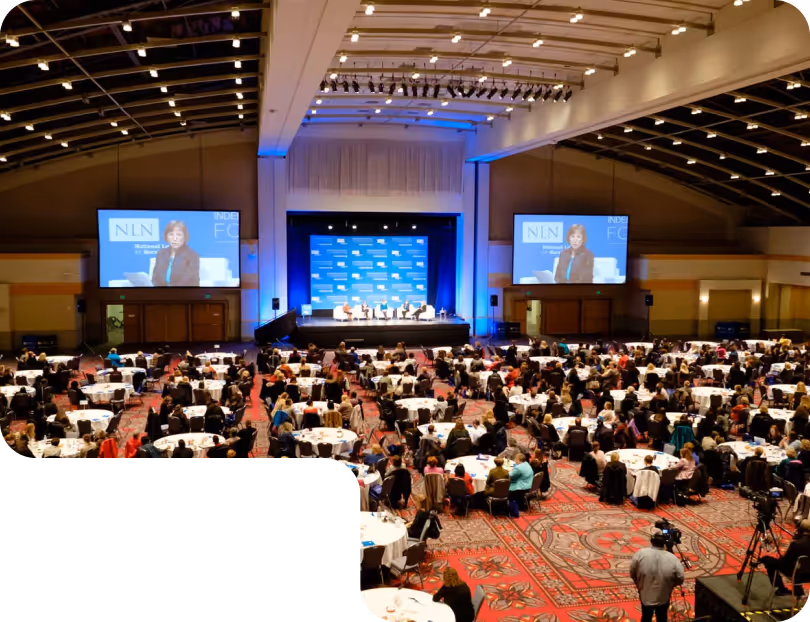 Large conference room with many attendees seated at round tables facing a stage with panelists and two large screens displaying a speaker.