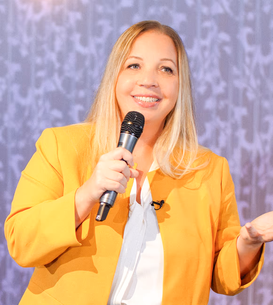Smiling woman with blonde hair in a yellow blazer holding a microphone, speaking on a stage with a patterned purple backdrop.