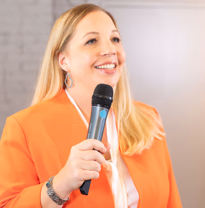 Smiling woman with long blonde hair wearing an orange blazer and holding a microphone.