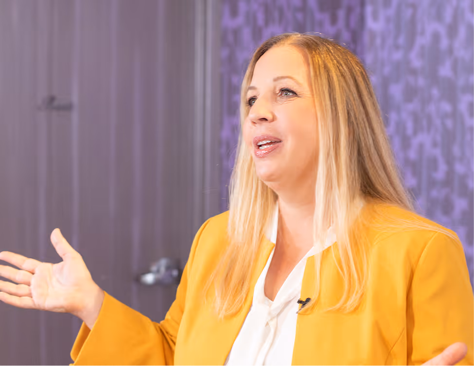 Woman with long blonde hair wearing a mustard yellow blazer and white blouse, gesturing with one hand while speaking.